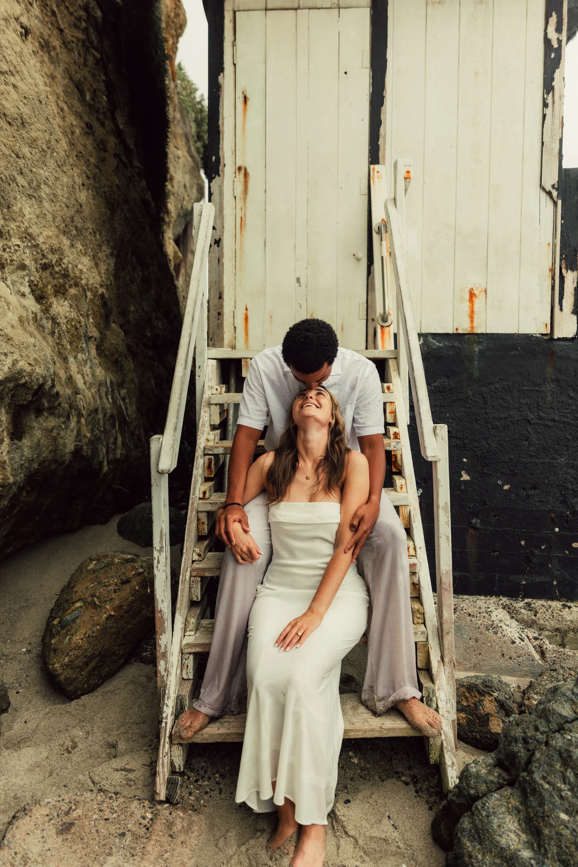 engagement photo outfits in Laguna Beach, couple in white dress and casual attire posing against a weathered coastal structure