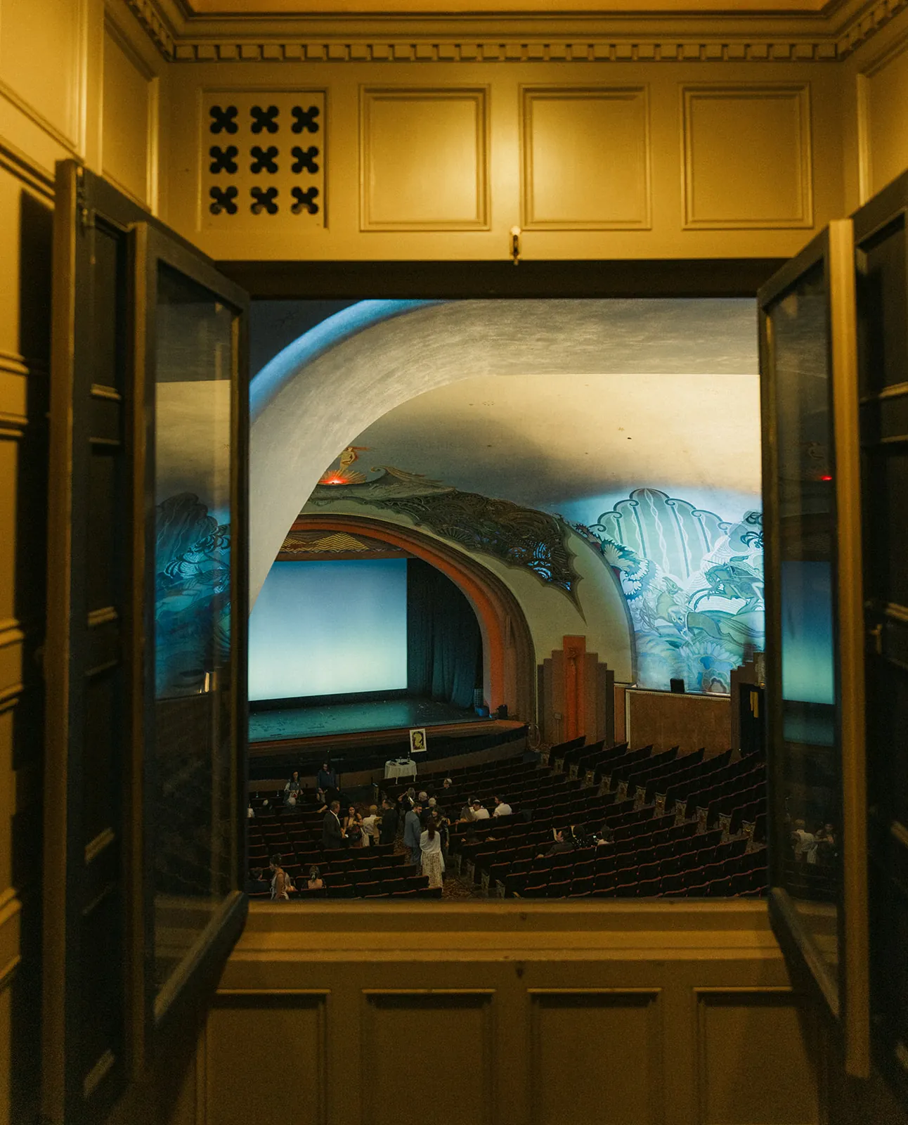 View through open doors into the Avalon Theatre at the Catalina Island Casino, showing the art deco interior, teal Beckman murals, and guests arriving for the wedding ceremony