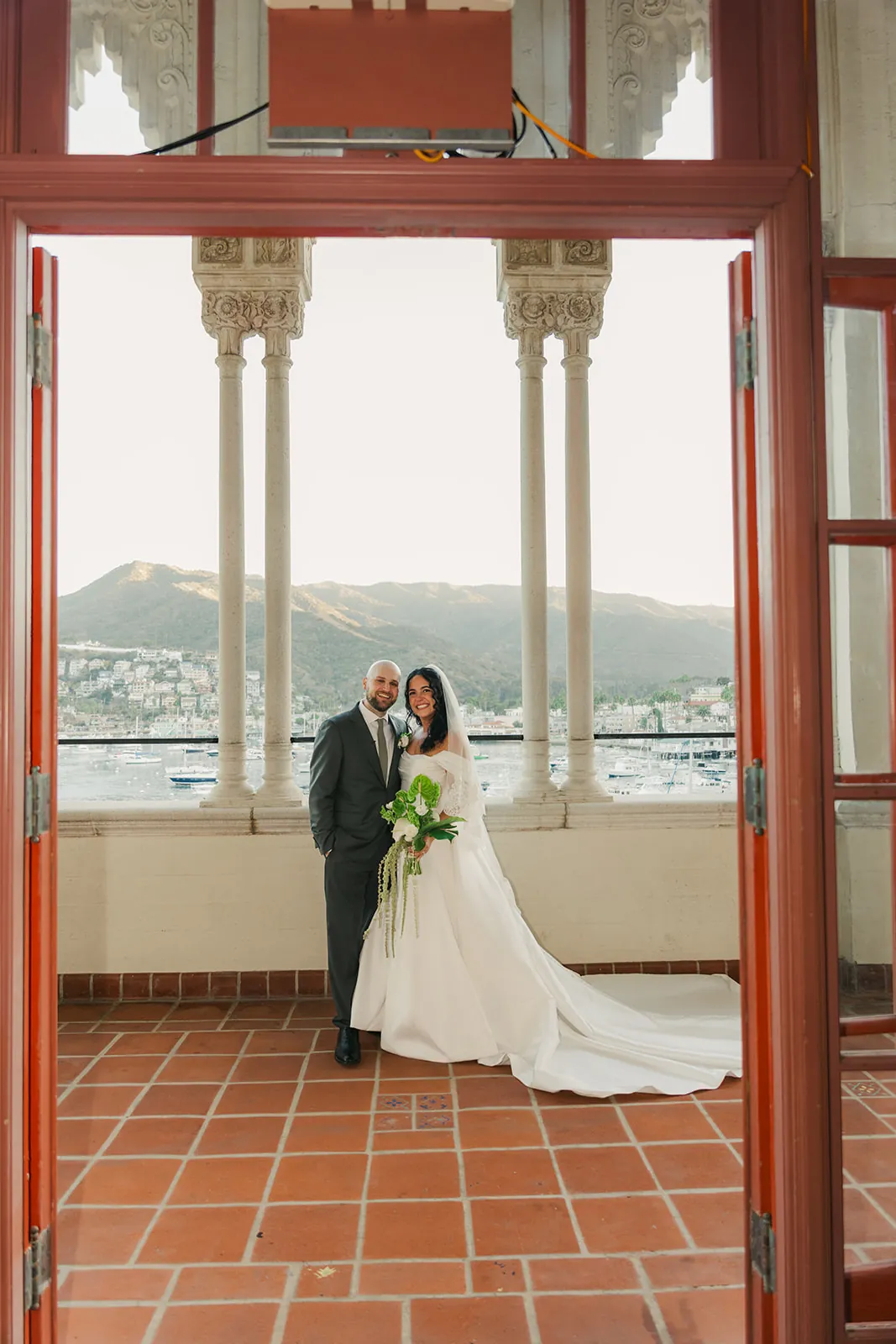Bride and groom standing in a red doorframe on the Casino colonnade veranda, white ornate columns and Avalon Bay behind them at their Catalina Island Casino wedding