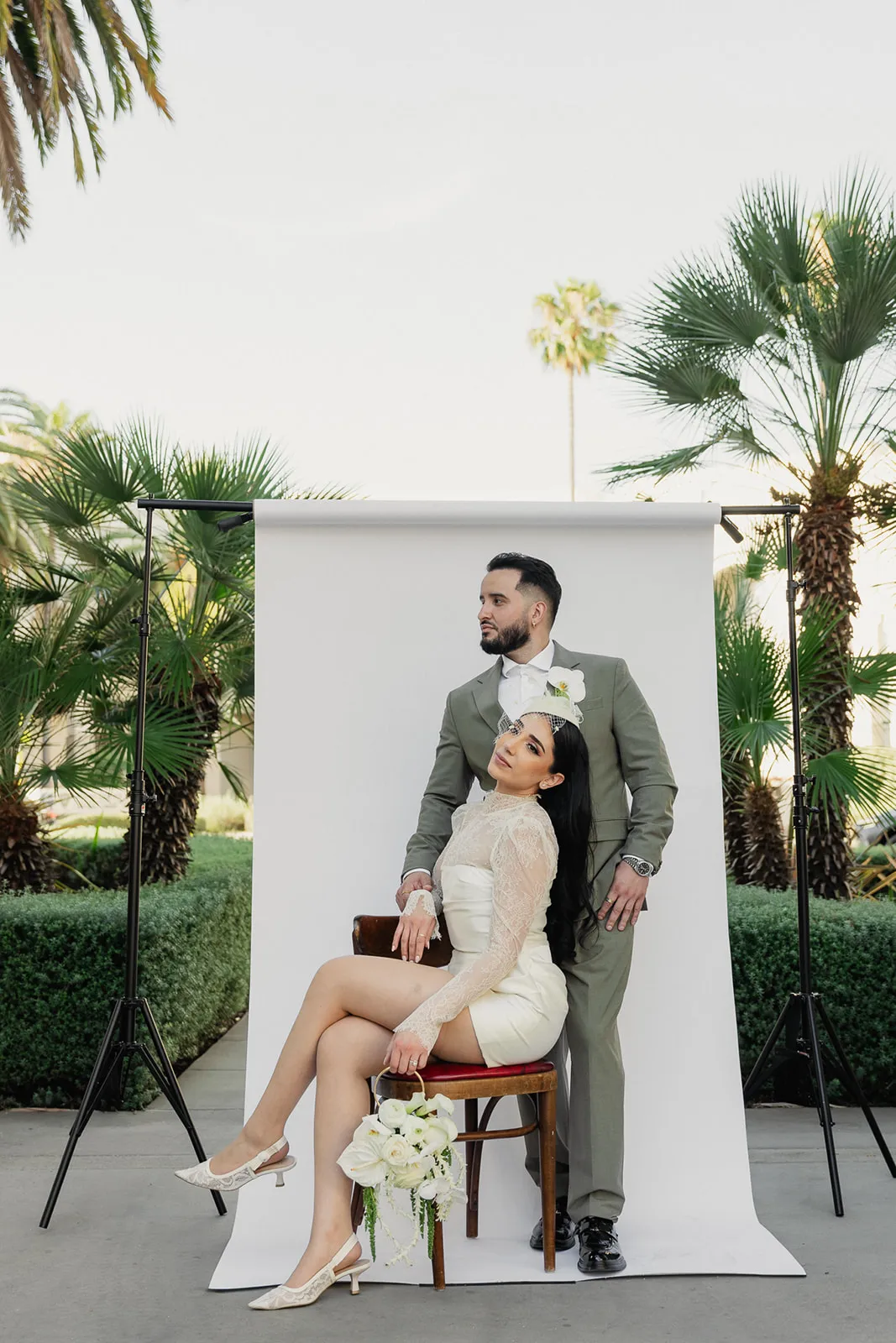 Editorial couple portrait against a white studio backdrop set up outdoors at Beverly Hills City Hall, bride seated on a bentwood stool in short lace wedding dress and birdcage fascinator with cascading white bouquet, groom standing behind her in olive double-breasted suit, palm trees and hedgerows behind the setup, photographed by Los Angeles wedding photographer Rene Porto