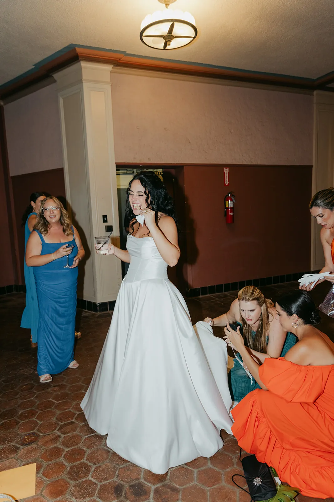 Bride eating during the Catalina Island Casino wedding reception while bridesmaids help with her dress in the corridor