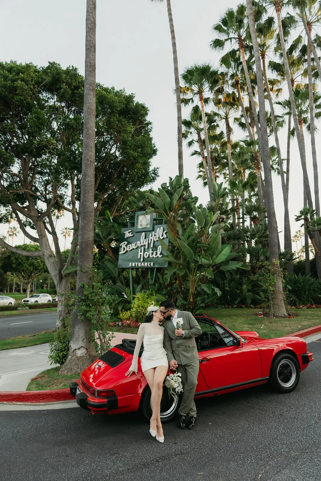 Couple kissing against a vintage red Porsche 911 in front of the Beverly Hills Hotel entrance sign after their courthouse wedding, bride in short white lace dress and fascinator, groom in olive double-breasted suit, Beverly Hills elopement photography by Rene Porto