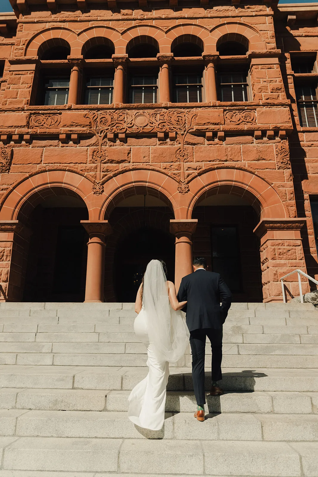 Couple walking up the red sandstone steps of the Old Orange County Courthouse toward the arched Romanesque facade on their wedding day