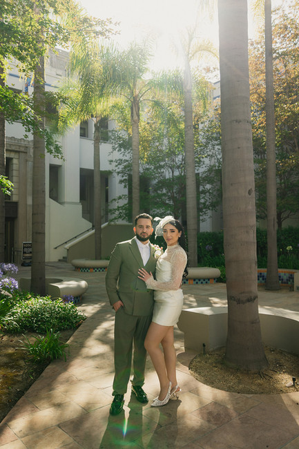 Couple portrait in the palm-lined courtyard at Beverly Hills City Hall with natural sun flare, bride in short lace wedding dress and fascinator leaning into groom in olive suit, photographed during a Beverly Hills courthouse elopement by Los Angeles wedding photographer Rene Porto