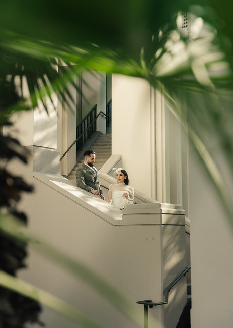 Couple on the exterior staircase of Beverly Hills City Hall framed through palm fronds, bride in short white lace turtleneck dress and birdcage fascinator holding white bouquet, groom in olive double-breasted suit, Beverly Hills courthouse wedding photography by Rene Porto