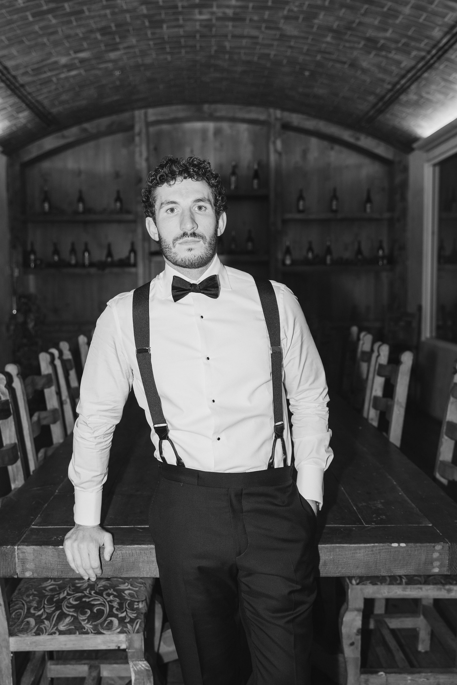 black and white portrait of the groom in suspenders standing inside the winery barrel room before the ceremony