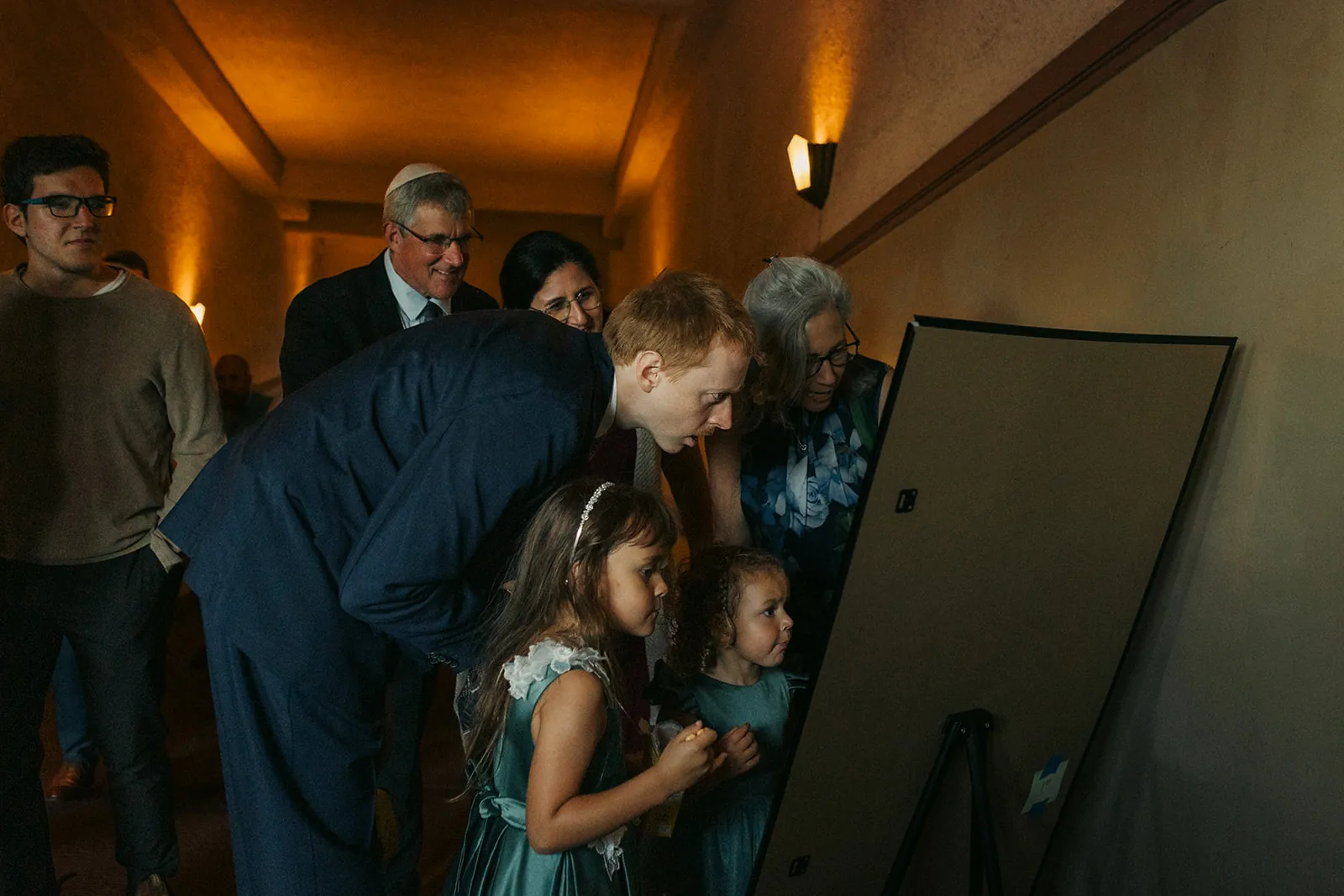 Wedding guests gathered around the couple's photo postboard in the Catalina Island Casino corridor during cocktail hour, candid documentary moment
