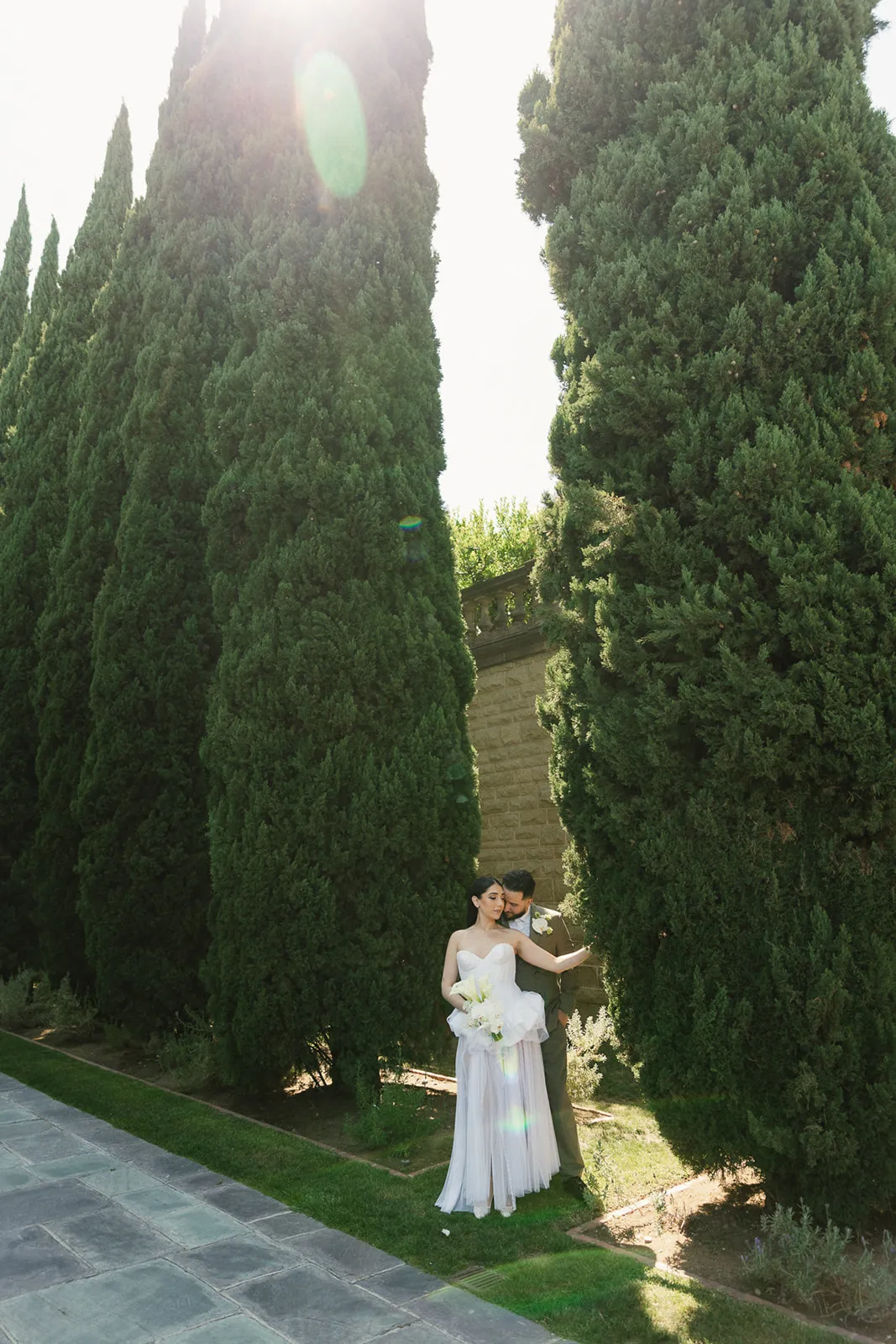 Couple embracing between tall Italian cypress trees at Greystone Mansion in Beverly Hills, bride in flowing strapless white chiffon gown with white bouquet, groom in olive suit, sun flare through the canopy above