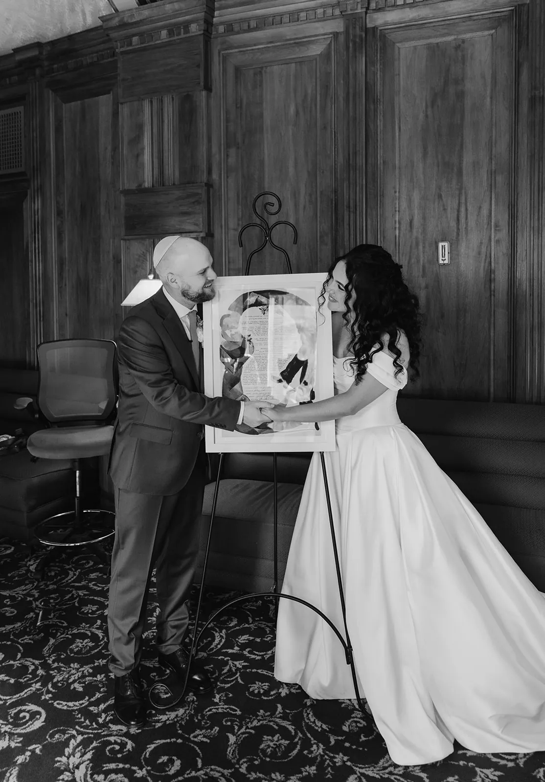 Bride and groom holding their custom ketubah art piece together in the dark wood-paneled room at the Catalina Island Casino, black and white