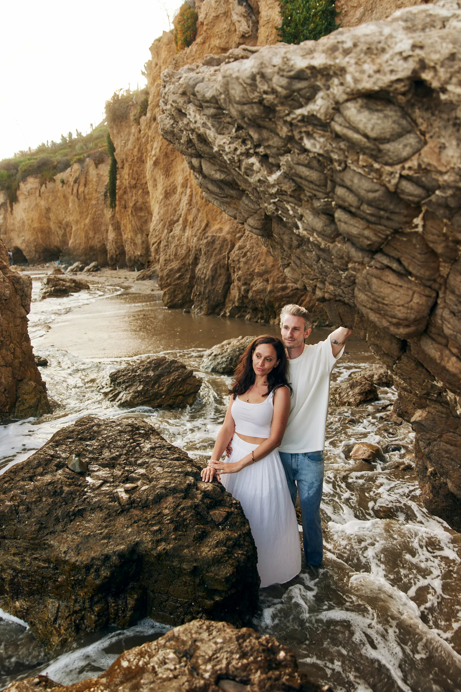 engagement photo outfits at El Matador Beach Malibu, couple in white crop top maxi skirt and casual white t-shirt posing on coastal rocks with ocean waves