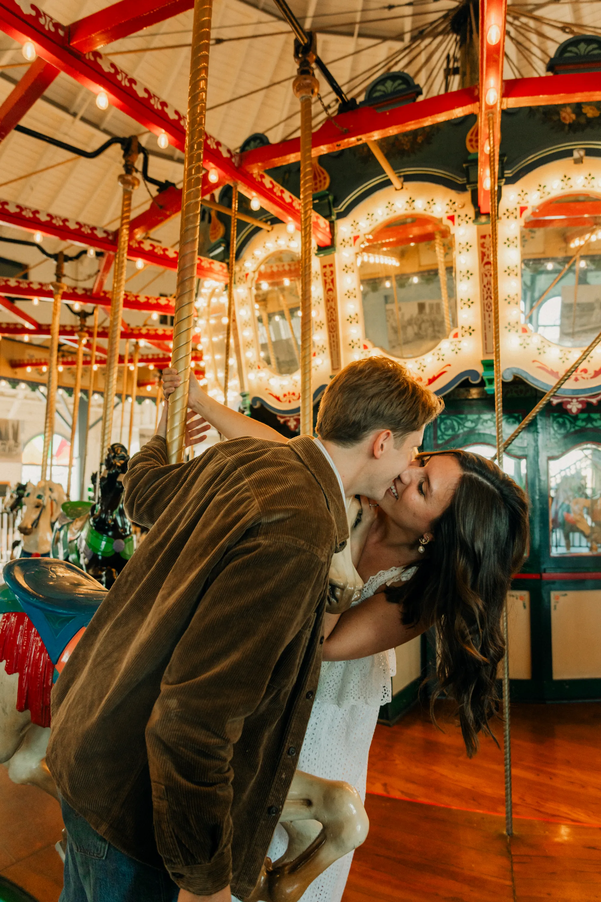 engagement photo outfits at Santa Monica Pier, couple in brown velvet jacket and white sundress sharing a kiss on the historic carousel