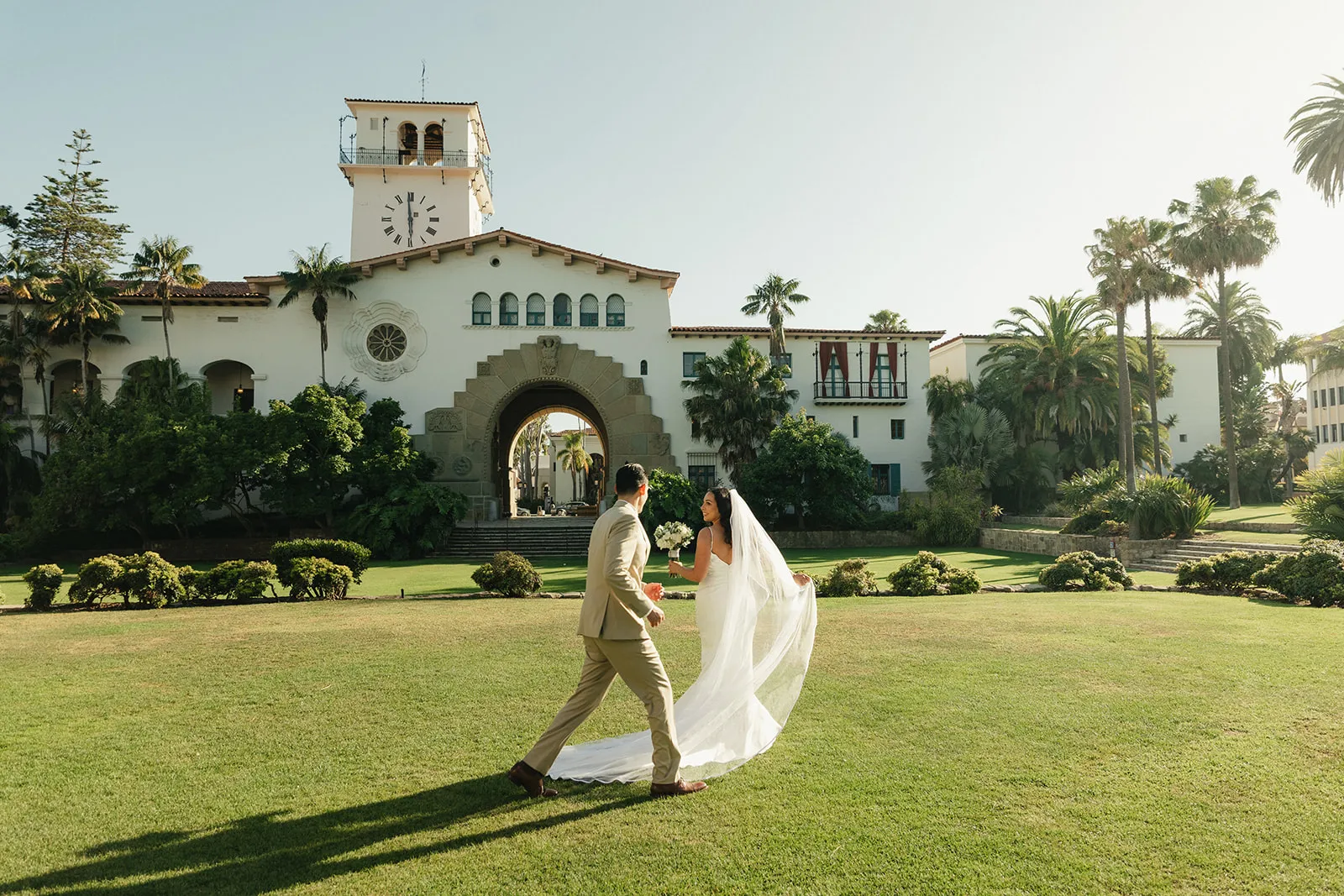 Wide editorial shot of bride in a flowing veil walking across the front lawn of the Santa Barbara Courthouse with the clock tower in the background