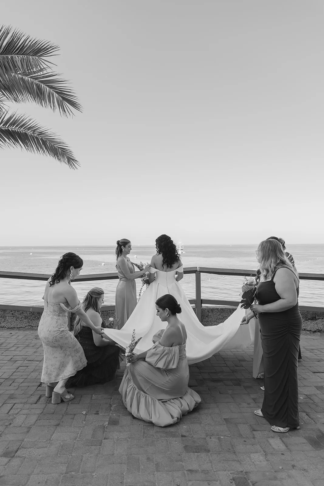 Bridesmaids kneeling to fix the bride's train along the Avalon waterfront before the Catalina Island Casino wedding ceremony, black and white