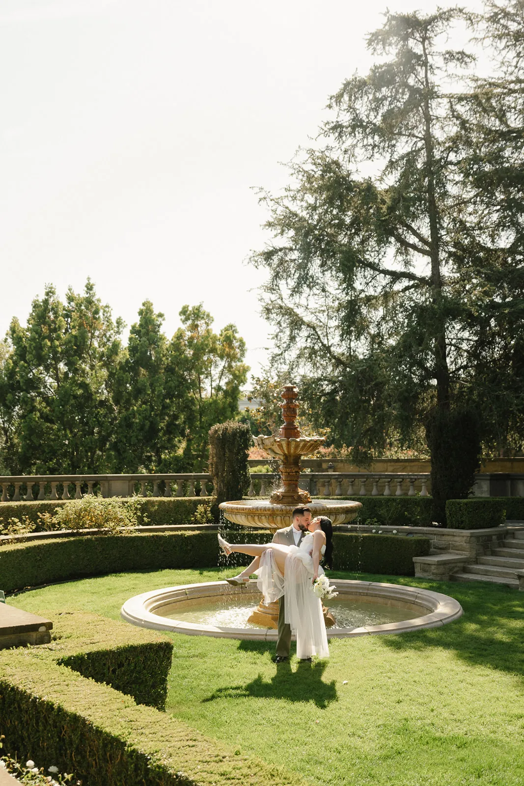 Groom lifting bride for a kiss at the ornate fountain courtyard at Greystone Mansion in Beverly Hills, bride in flowing white chiffon gown, formal gardens and stone balustrade in the background, Beverly Hills courthouse wedding photography by Rene Porto