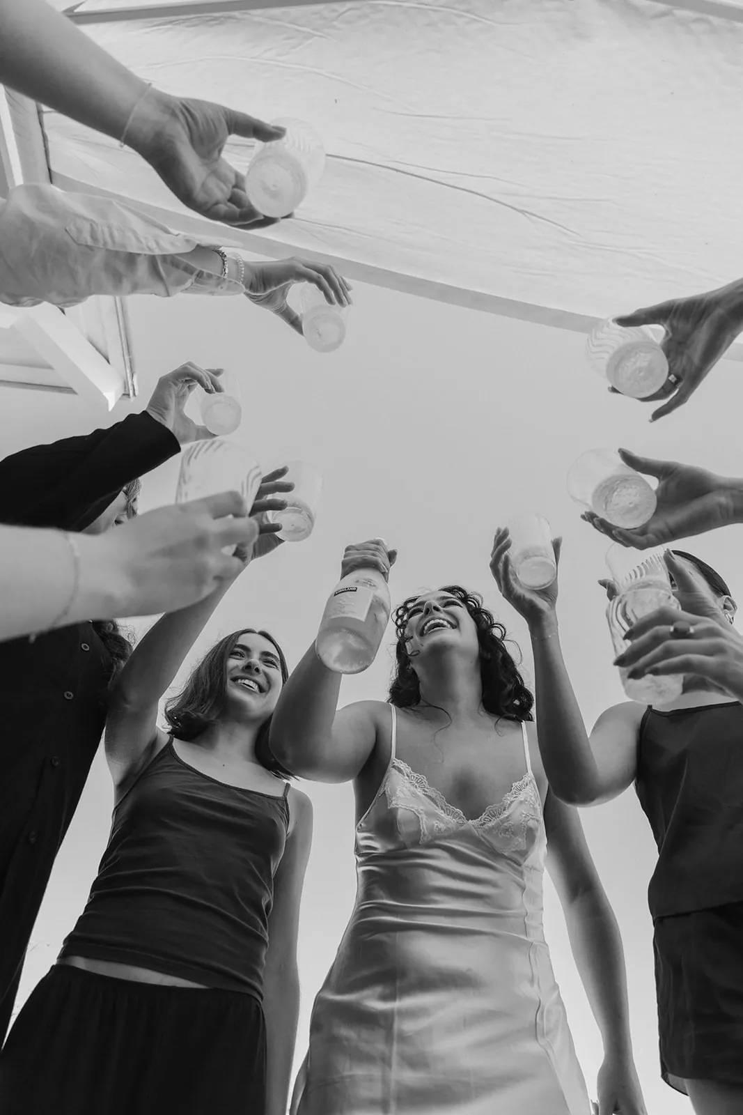 Bride and bridesmaids raising their arms in a celebratory toast during getting ready at their Catalina Island Casino wedding, photographed from below in black and white