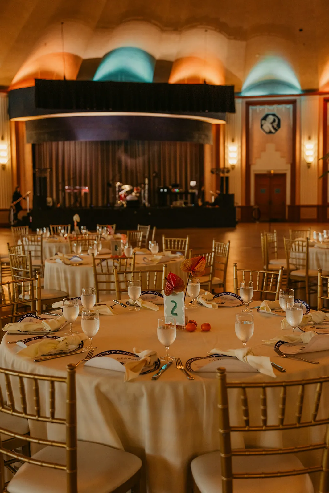 Catalina Island Casino Ballroom set for the wedding reception with round tables, chiavari chairs, and the stage visible under amber and teal uplighting