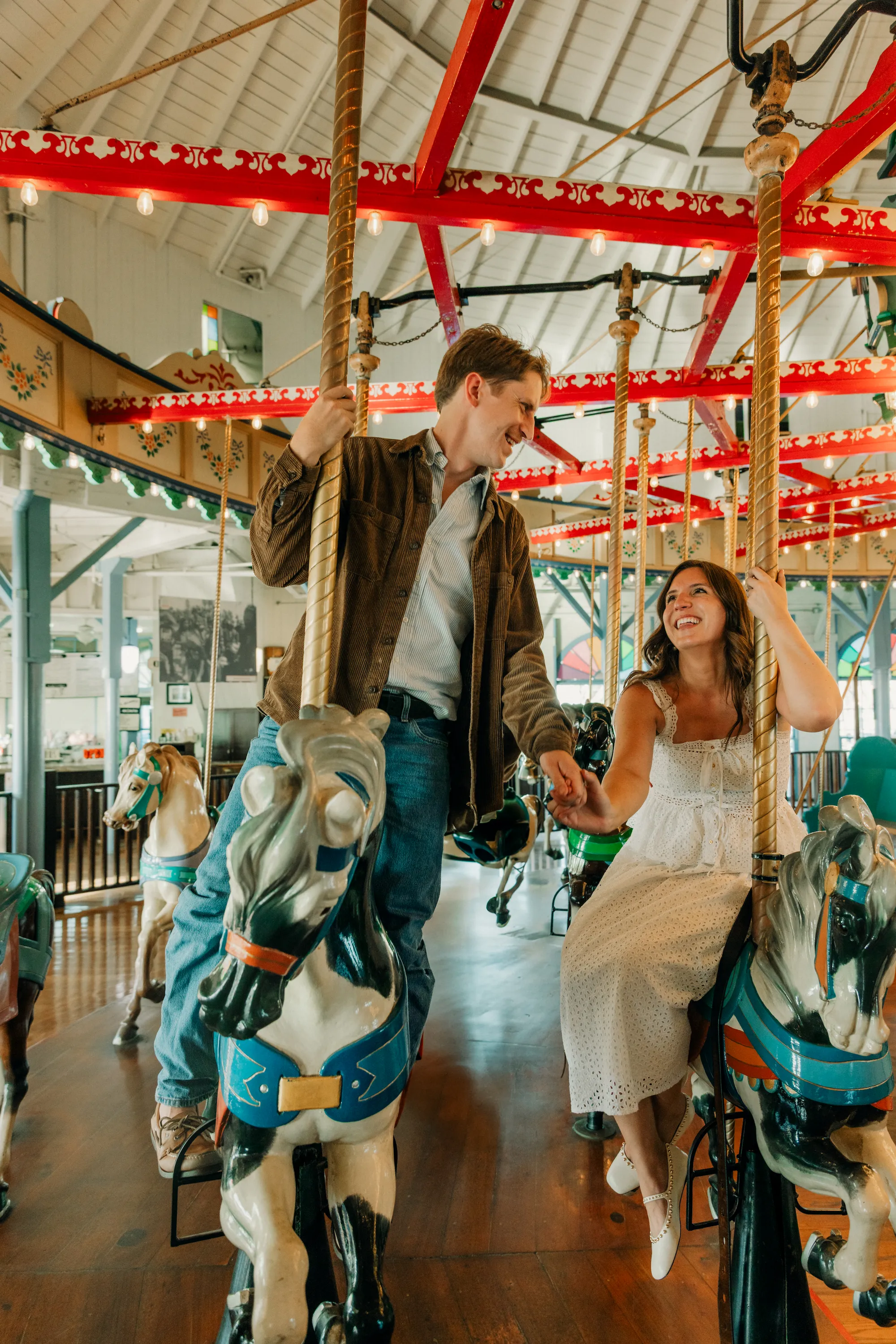 engagement photo outfits at Santa Monica Pier, couple in brown corduroy jacket and white eyelet dress laughing together on the Santa Monica carousel
