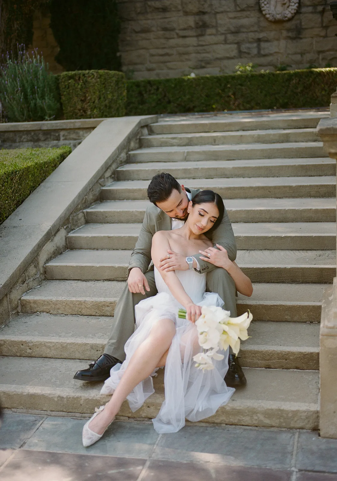 Intimate portrait of couple seated on the stone garden stairs at Greystone Mansion in Beverly Hills, groom in olive suit embracing bride from behind, bride in strapless white chiffon gown holding white calla lily bouquet, Beverly Hills courthouse wedding photography by Rene Porto