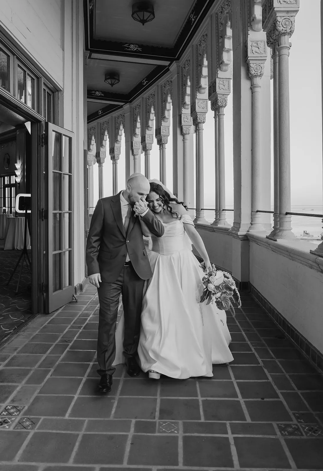 Groom kissing bride's hand as they walk laughing through the Casino colonnade, ocean visible through the arched columns at their Catalina Island wedding
