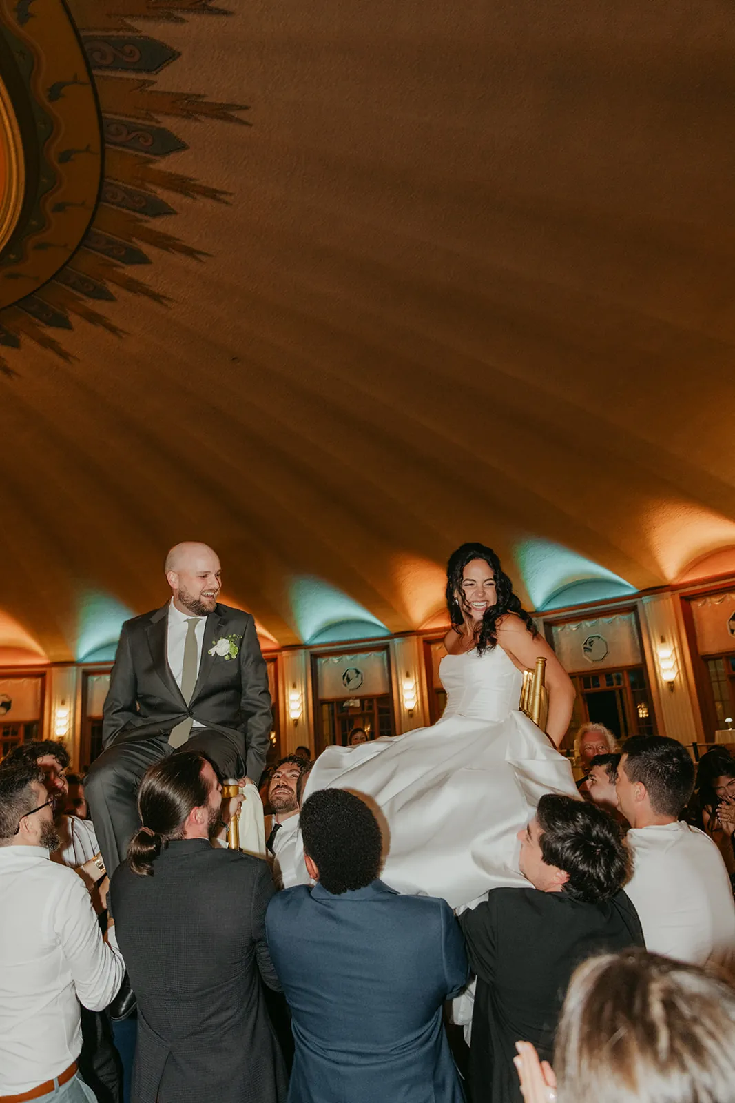 Bride and groom lifted on chairs by guests during the hora at their Catalina Island Casino Ballroom wedding reception, art deco ceiling and teal uplighting above