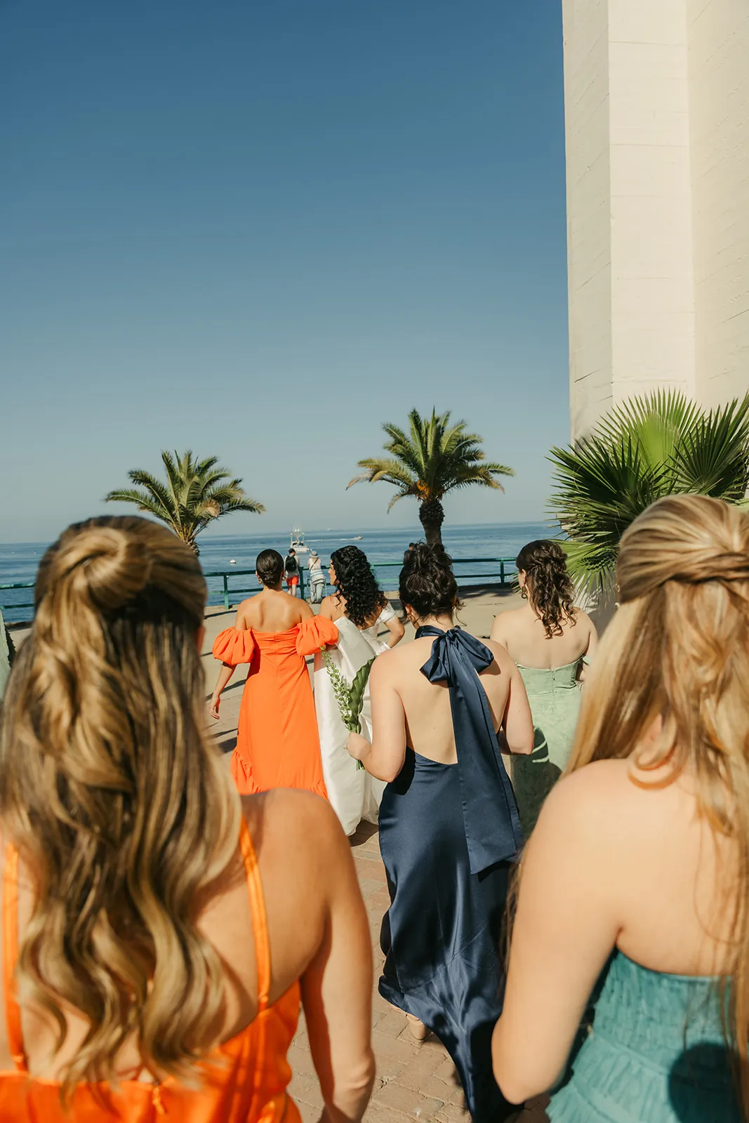 Bridal party in colorful dresses walking toward the Catalina Island Casino with palm trees and the ocean ahead, backs to camera