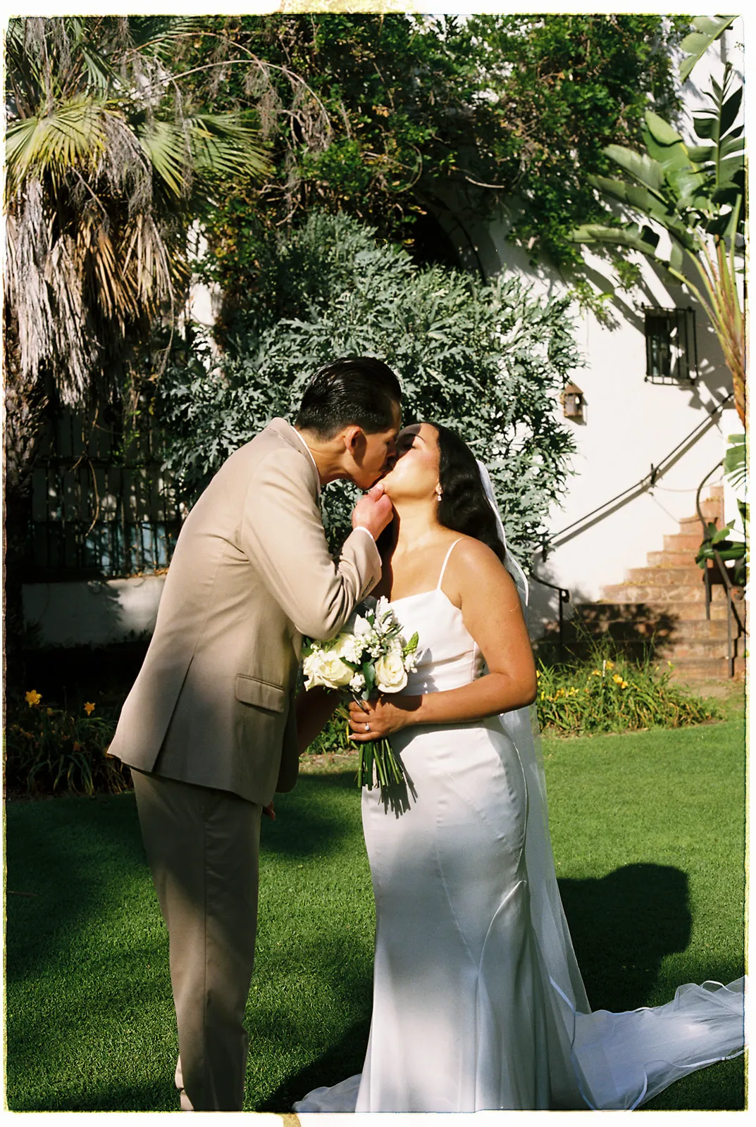 Couple kissing on the green lawn at the Santa Barbara Courthouse, groom in tan suit, bride holding a white bouquet