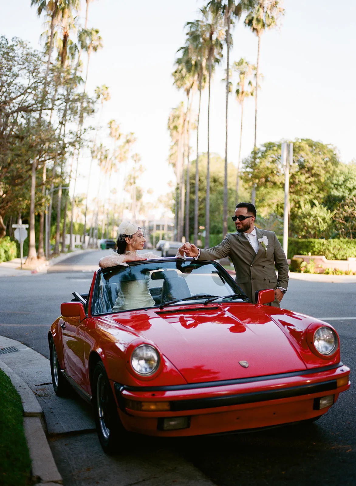 Couple sharing a playful fist bump across a vintage red Porsche 911 on a palm-lined Beverly Hills street at golden hour, bride in lace dress and fascinator inside the convertible, groom in olive suit and sunglasses outside, shot on film