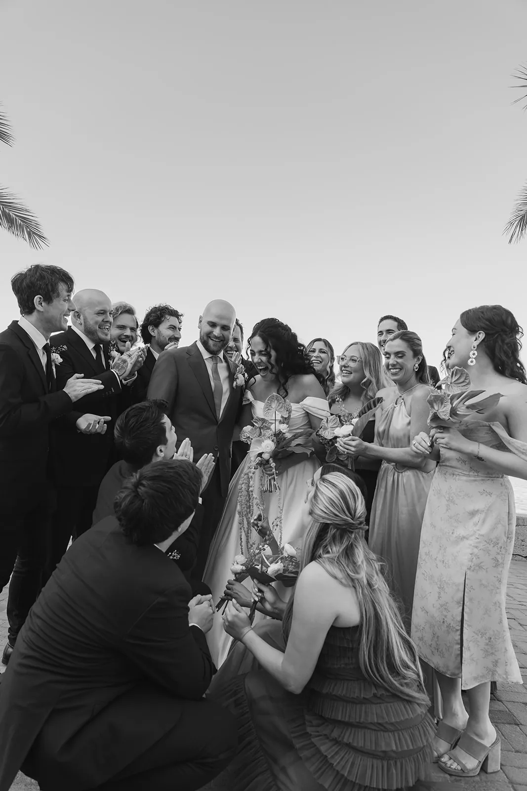 Full bridal party laughing together around the bride and groom outside the Catalina Island Casino, captured in black and white with palm trees framing the scene