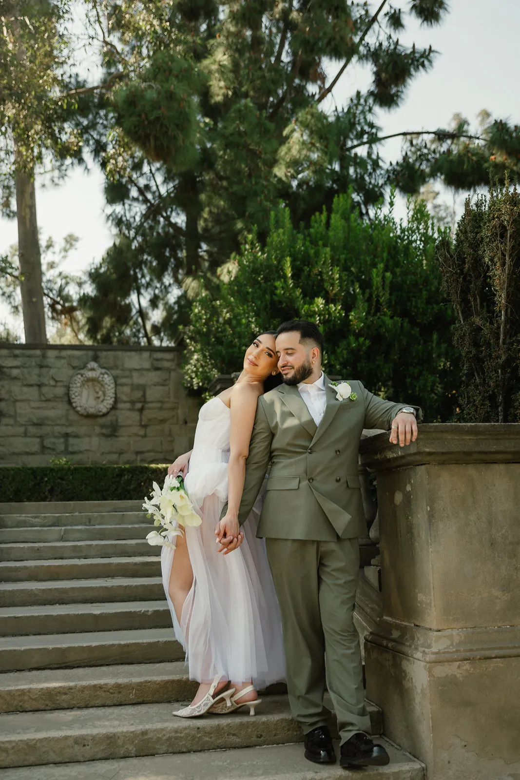Couple leaning together on the stone staircase at Greystone Mansion in Beverly Hills, bride in flowing strapless white chiffon dress resting her head on groom's shoulder, groom in olive double-breasted suit, formal garden and stone wall behind them