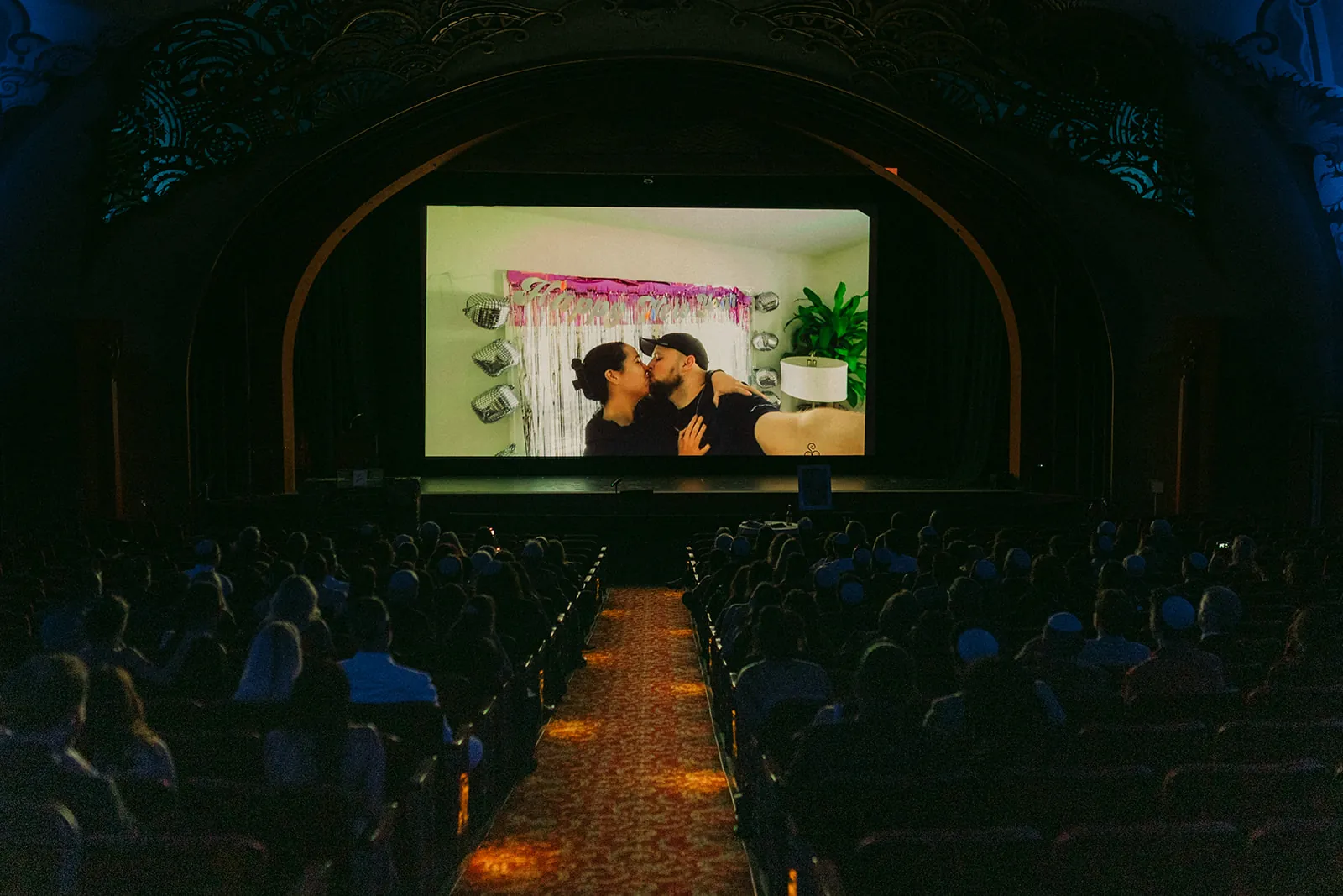 Sydney and Jason's love story video playing on the screen of the Avalon Theatre at the Catalina Island Casino during their wedding ceremony, guests seated in the art deco theatre watching from the audience