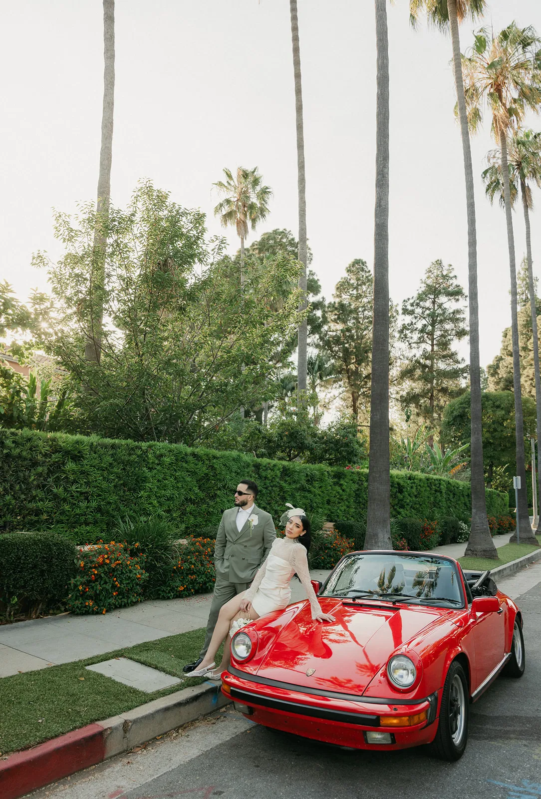 Bride sitting on the hood of a vintage red Porsche 911 on a palm-lined Beverly Hills street at sunset, groom standing beside her in olive suit and sunglasses, lush hedgerows and warm evening light behind them, photographed by Los Angeles wedding photographer Rene Porto