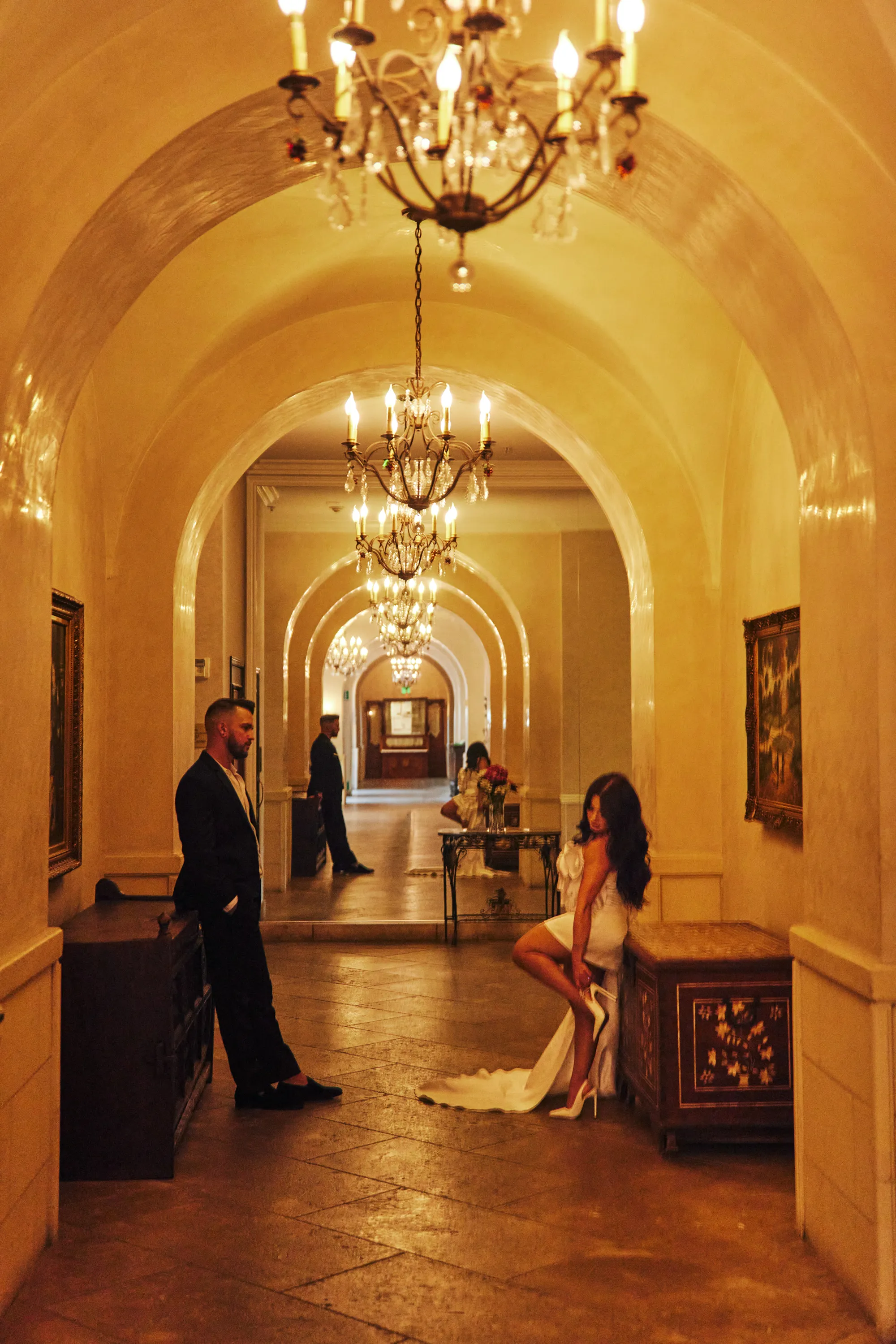 engagement photo outfits in a hotel lobby in Los Angeles, couple in black suit and white mini dress in a warm golden chandelier hallway
