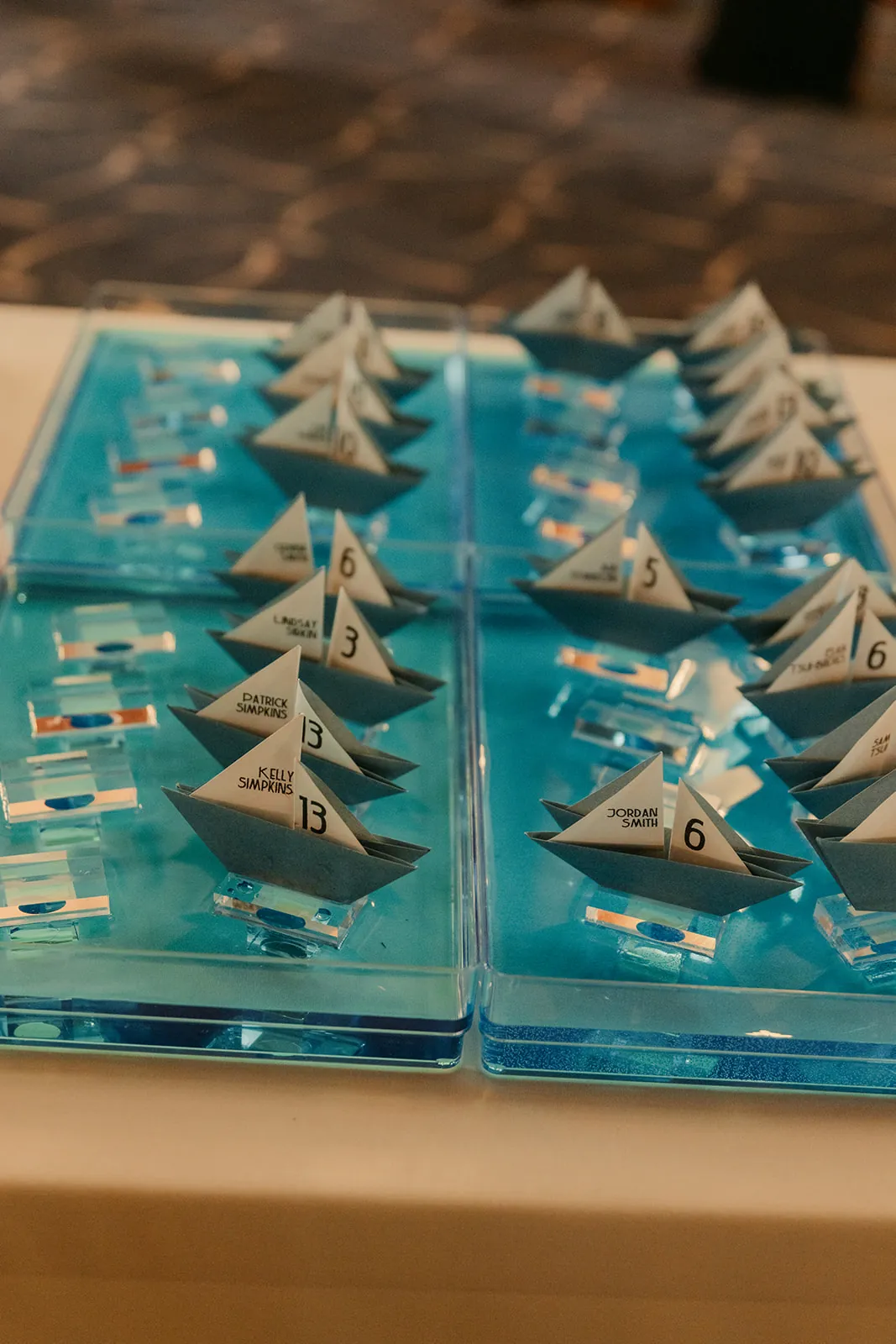 Custom origami sailboat escort cards arranged on blue acrylic trays at a Catalina Island Casino wedding reception, each boat printed with a guest name and table number