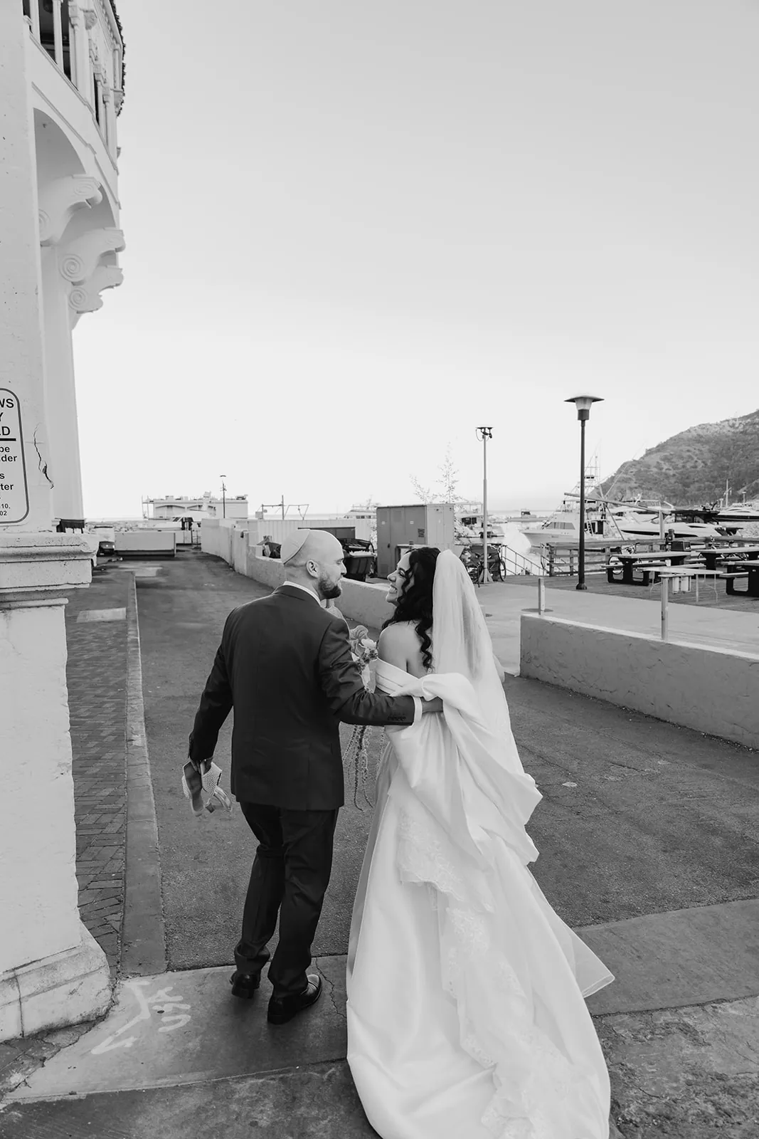 Newlyweds walking together outside the Catalina Island Casino along the harbor after their wedding ceremony, black and white