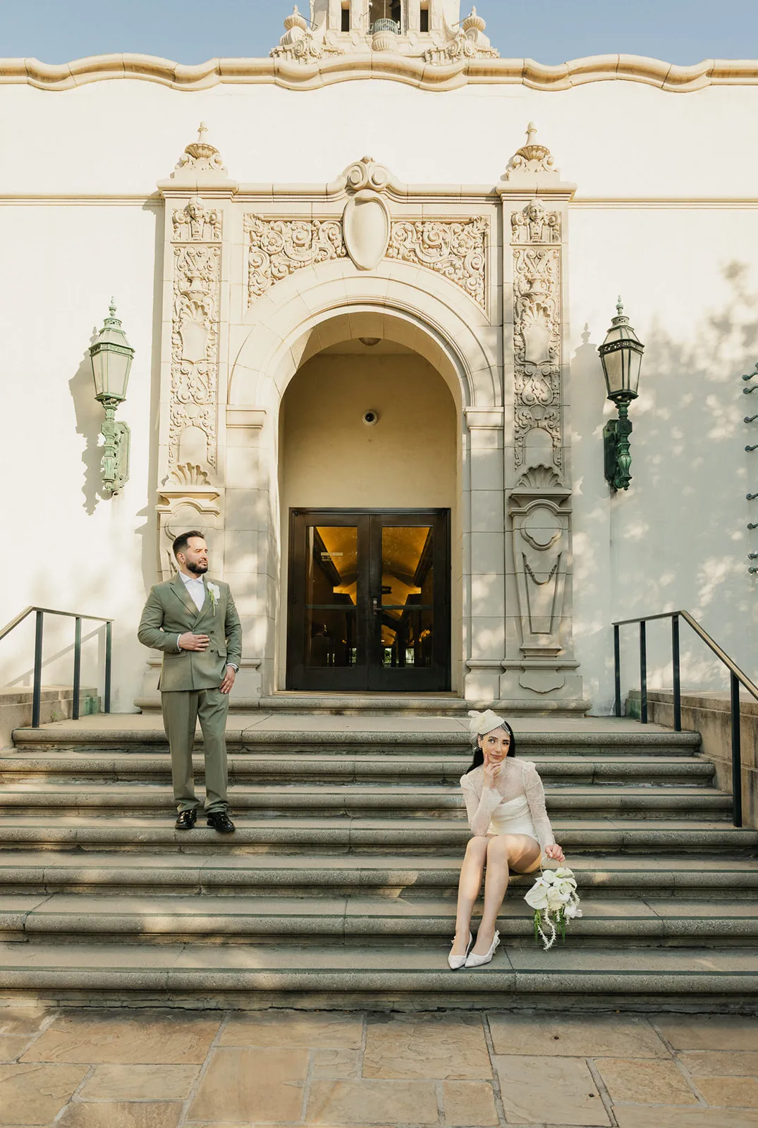 Couple on the steps of Beverly Hills City Hall during a courthouse wedding, bride in short lace dress and birdcage fascinator seated on steps with white bouquet, groom in olive double-breasted suit standing at the entrance arch, photographed by Los Angeles wedding photographer Rene Porto