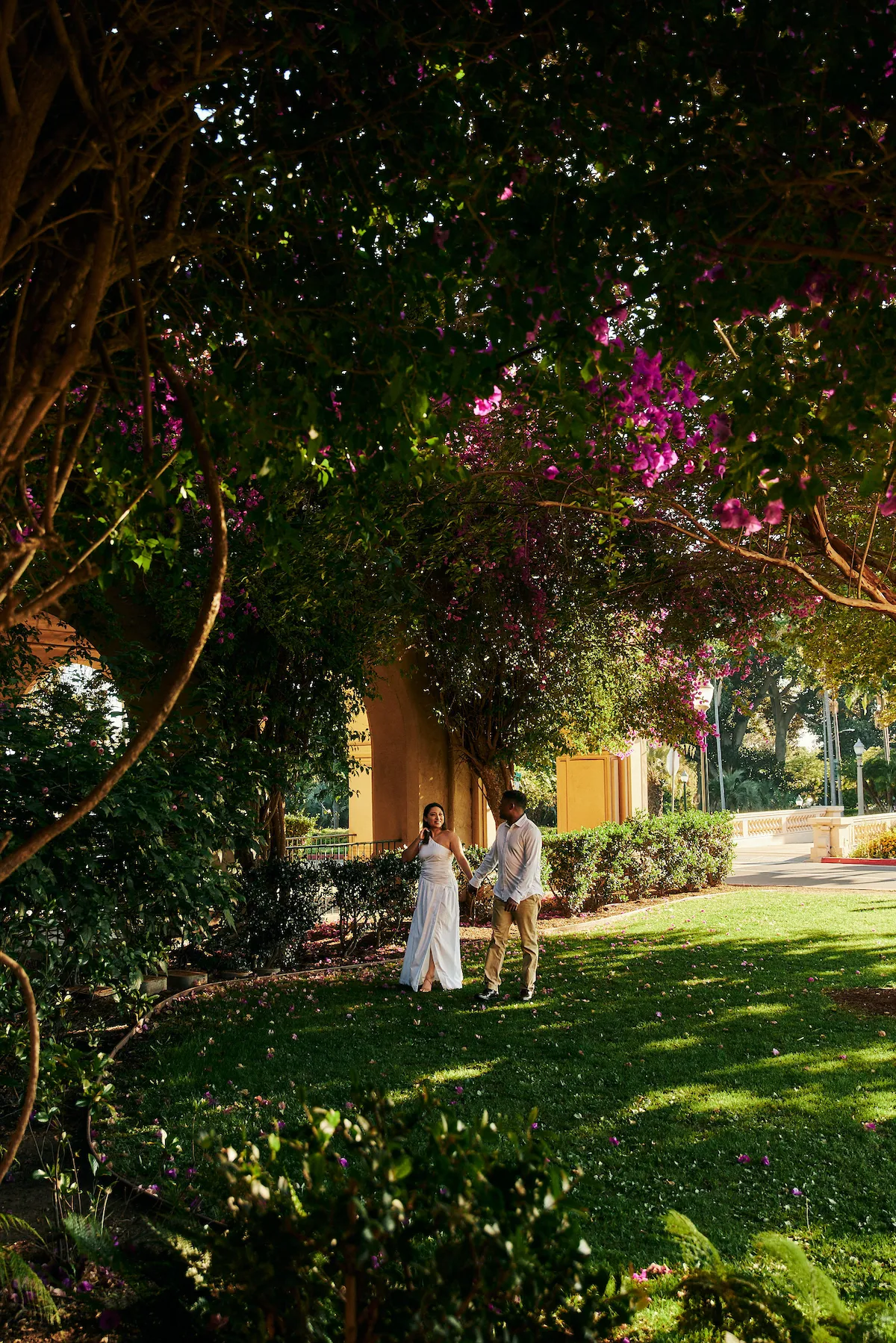 engagement photo outfits at Balboa Park San Diego, couple in white maxi dress and tan trousers walking hand in hand through a lush garden with blooming bougainvillea