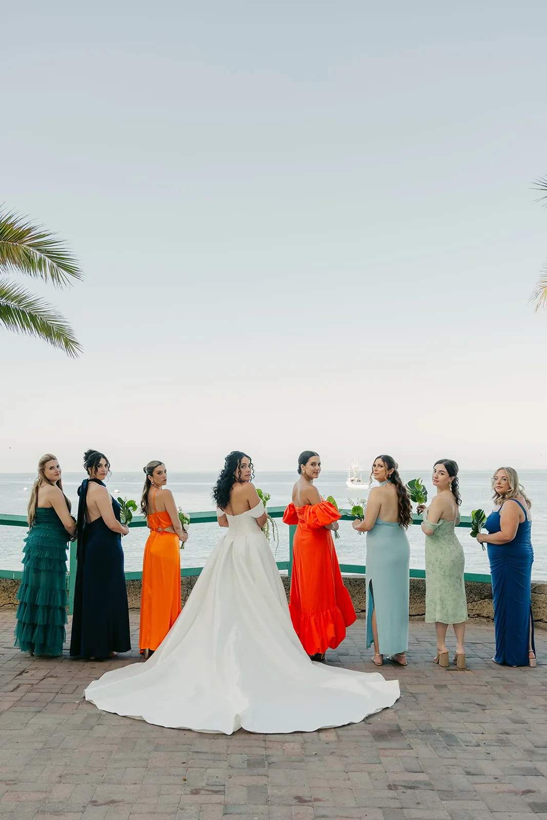 Bride and bridesmaids in colorful gowns looking back over their shoulders with Avalon Bay and a sailboat behind them at their Catalina Island destination wedding