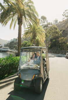 Wedding couple riding a golf cart through Avalon on Catalina Island, palm trees and the harbor visible along the street on their wedding day