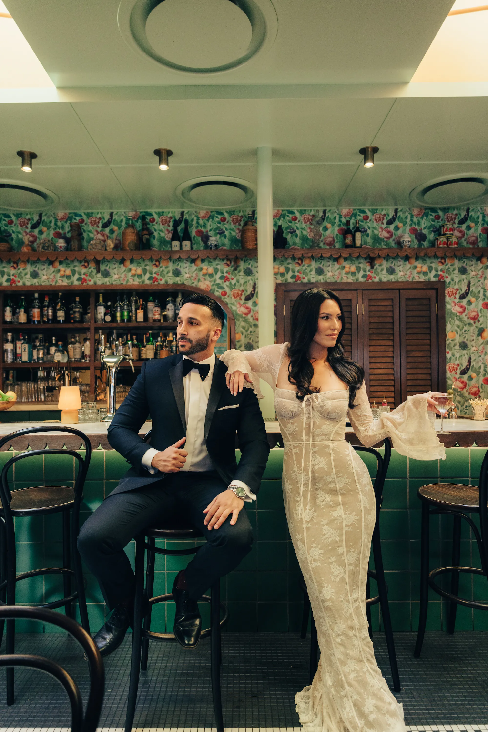 engagement photo outfits at a Los Angeles restaurant, couple in tuxedo and white lace gown seated at a moody bar with floral wallpaper