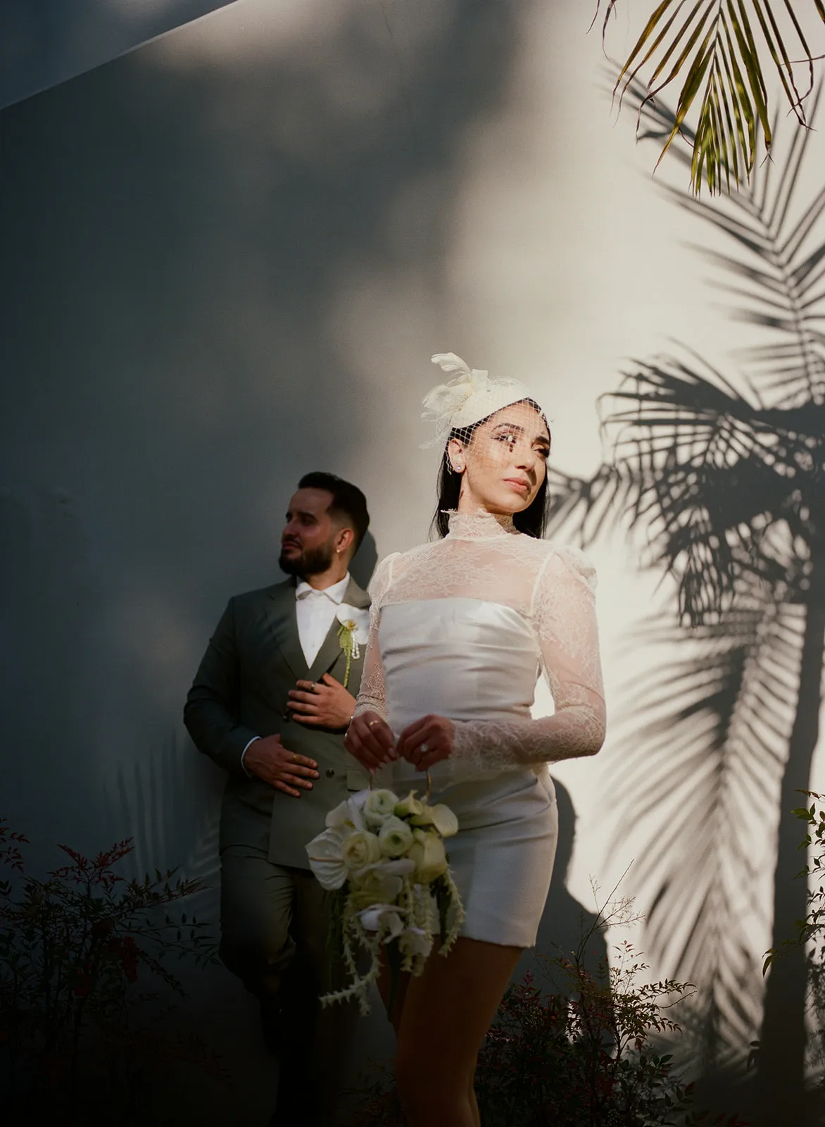 Moody editorial portrait of couple against a white wall with dramatic palm tree shadows at Beverly Hills City Hall, bride in short lace turtleneck wedding dress and birdcage fascinator in the foreground, groom in olive suit behind her, Beverly Hills courthouse wedding photography by Rene Porto
