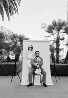 Black and white editorial portrait of couple against a white seamless studio backdrop at Beverly Hills City Hall, both wearing sunglasses, bride in short lace turtleneck dress and fascinator standing behind seated groom in olive suit, palm trees flanking both sides