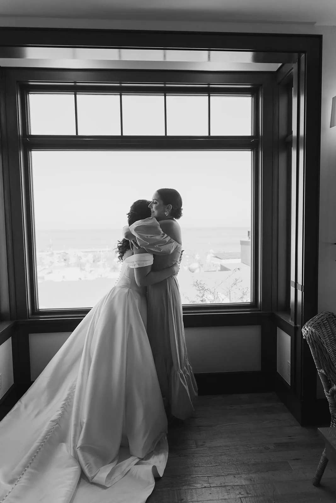Bride embracing a bridesmaid by a large window overlooking Avalon during getting ready at their Catalina Island Casino wedding, captured in black and white