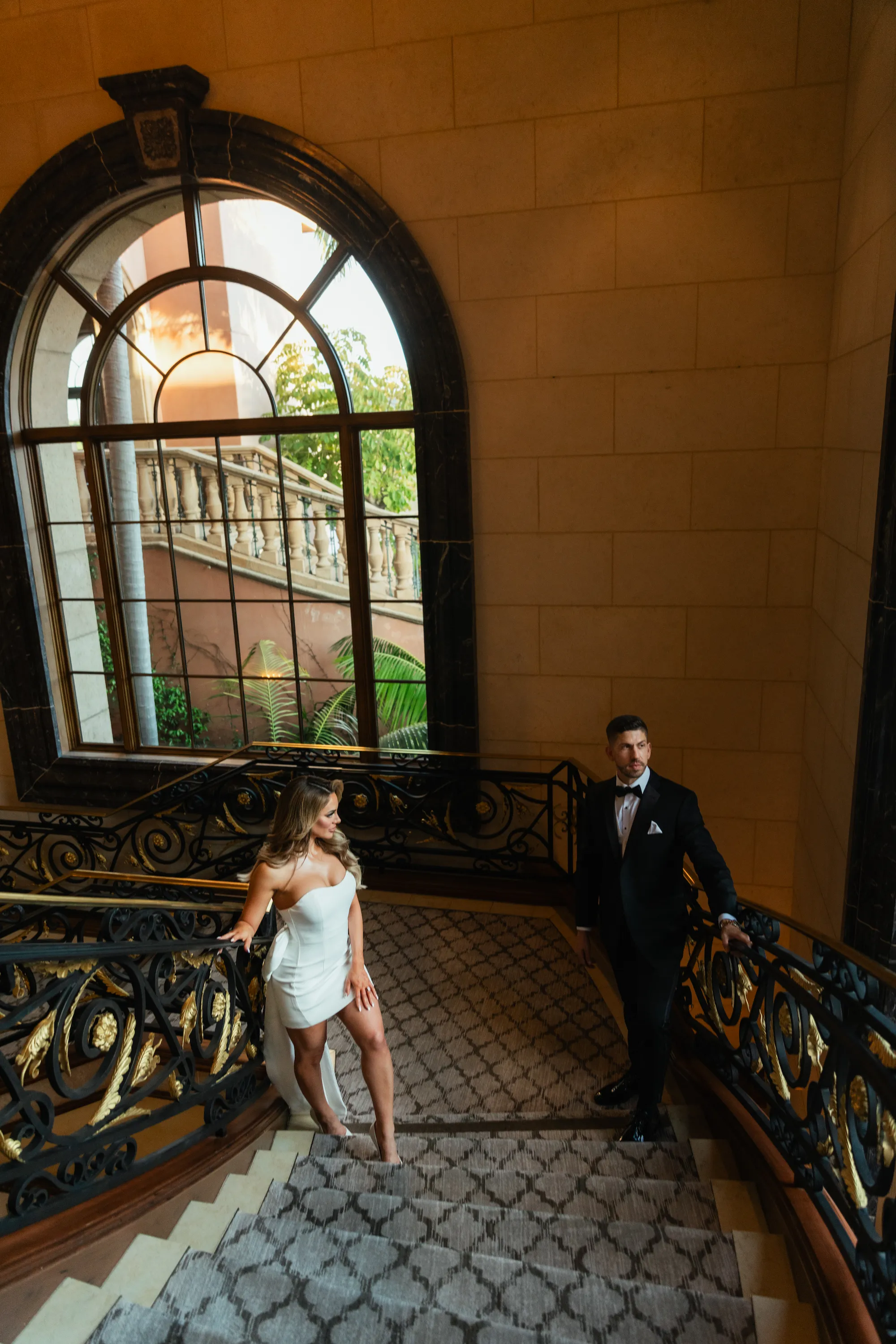 engagement photo outfits at Fairmont Grand Del Mar San Diego, couple in white mini dress and black tuxedo on an ornate gold iron staircase with a large arched window