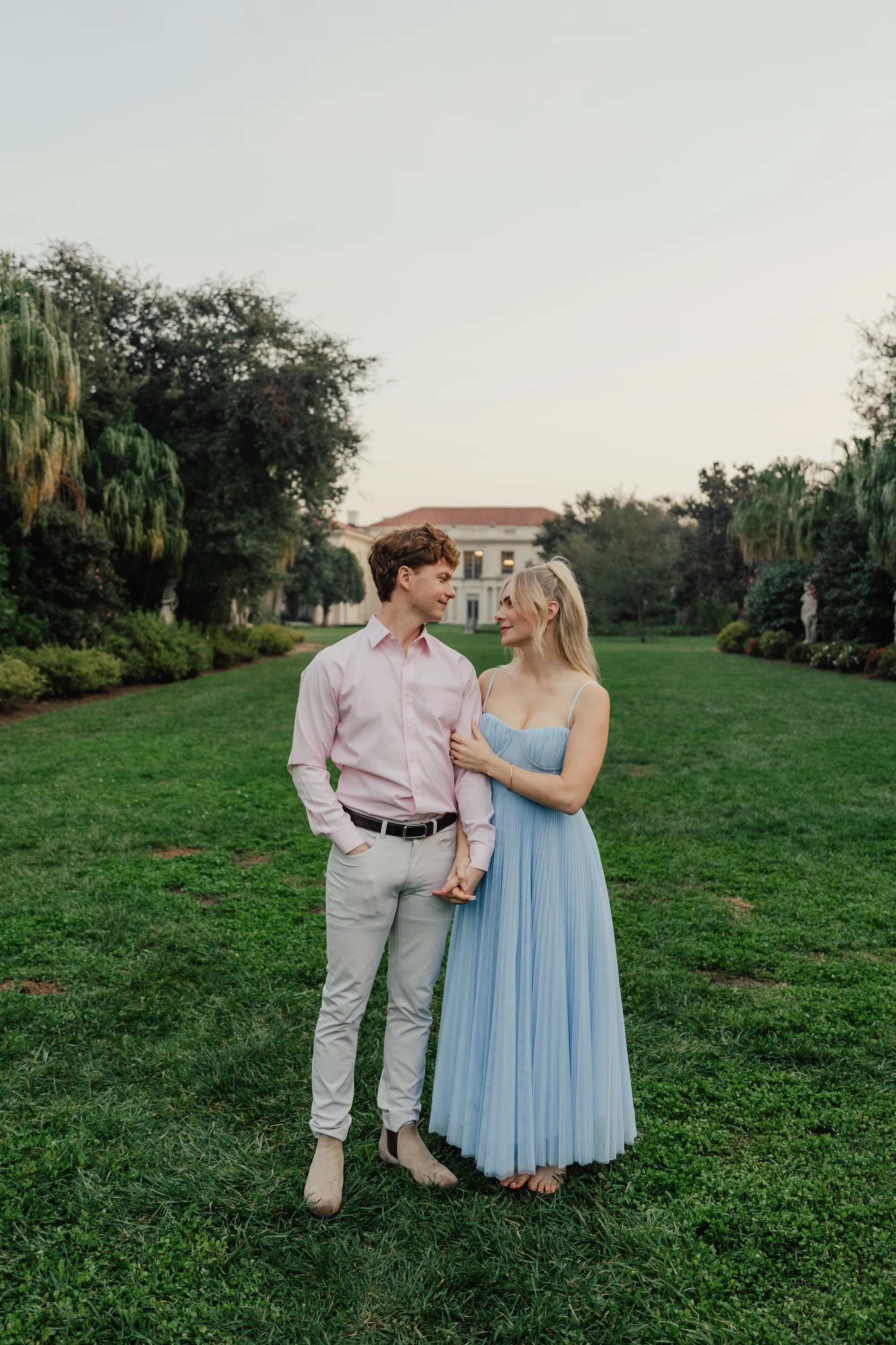 engagement photo outfits at the Huntington Library San Marino, couple in dusty blue pleated dress and pink shirt standing on the garden lawn with the estate in the background