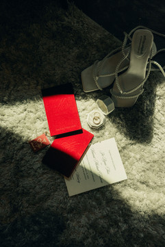Wedding details flat lay with red ring boxes, invitation suite, and pearl heels
