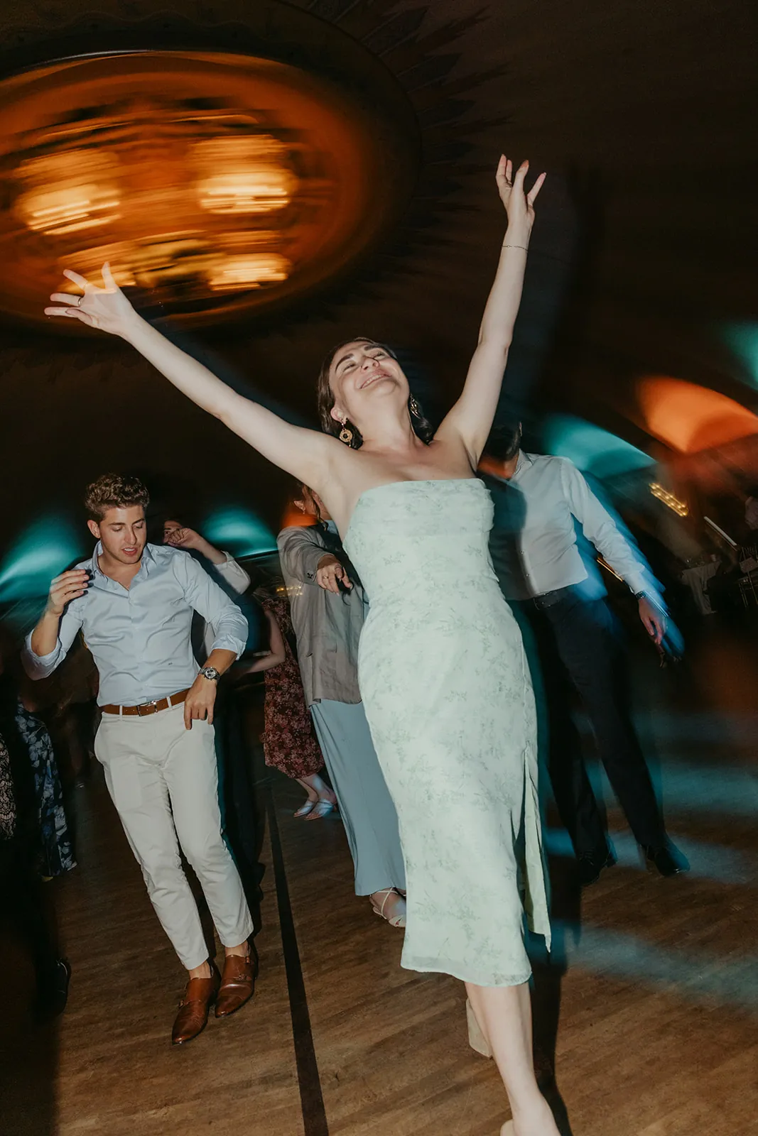 Guest dancing with arms raised on the Catalina Island Casino Ballroom floor, spinning ceiling detail and teal uplighting in motion blur