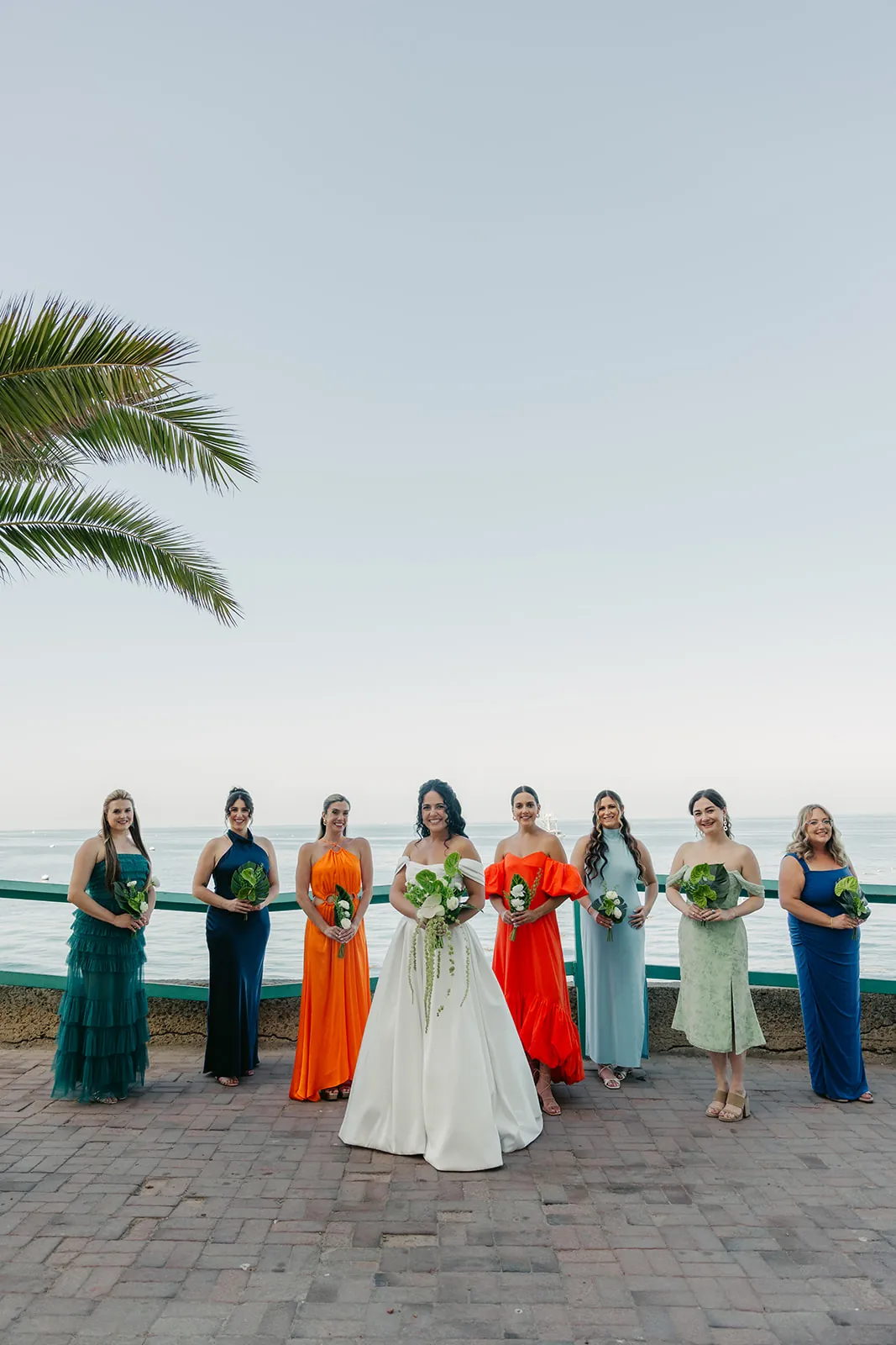 Full bridesmaids lineup in vibrant multicolor gowns along the Avalon waterfront at a Catalina Island Casino wedding, palm trees and ocean behind them