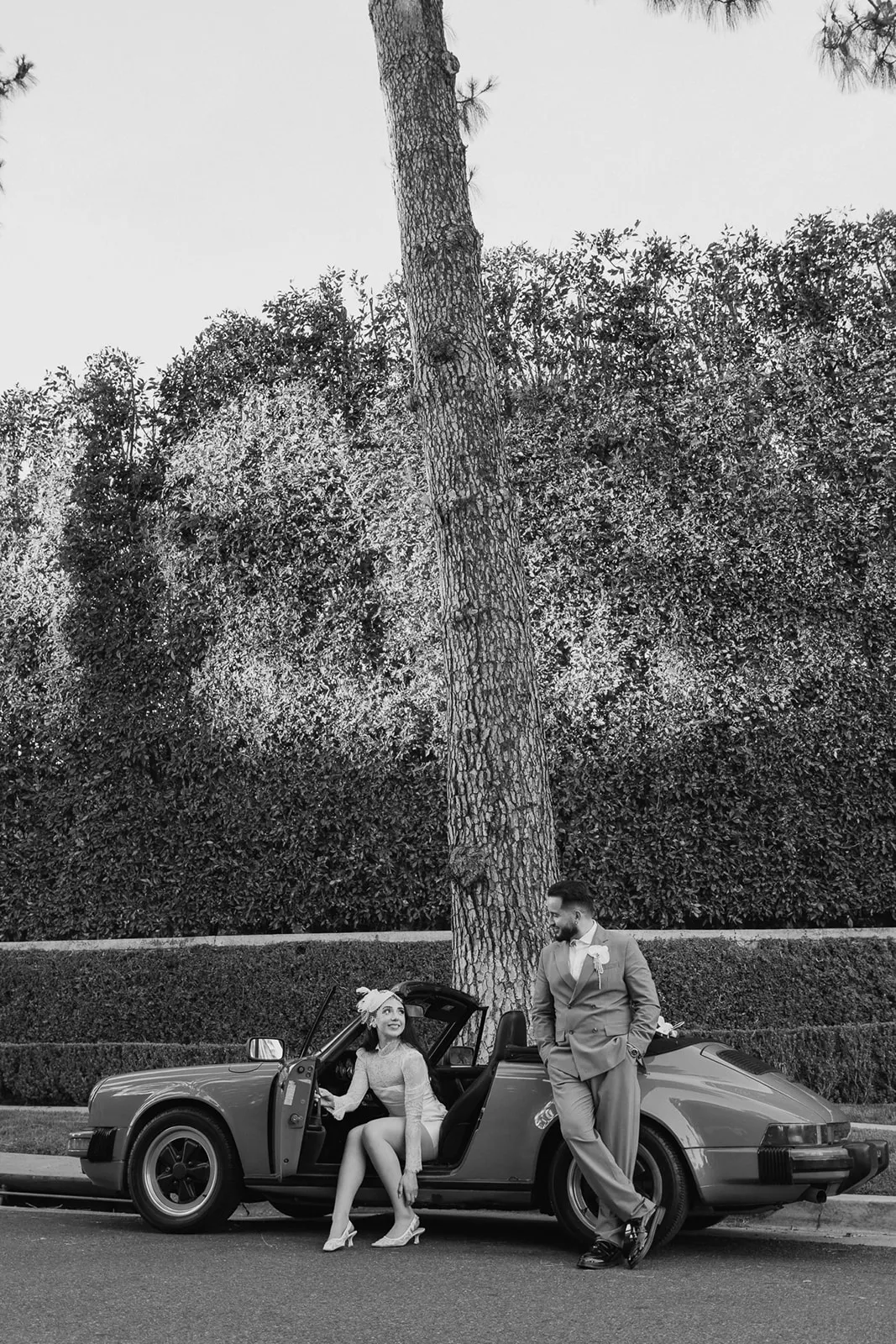 Black and white portrait of bride sitting in the open door of a vintage Porsche 911 while groom leans against the trunk in front of a Beverly Hills hedgerow, bride in short lace dress and fascinator, groom in double-breasted suit