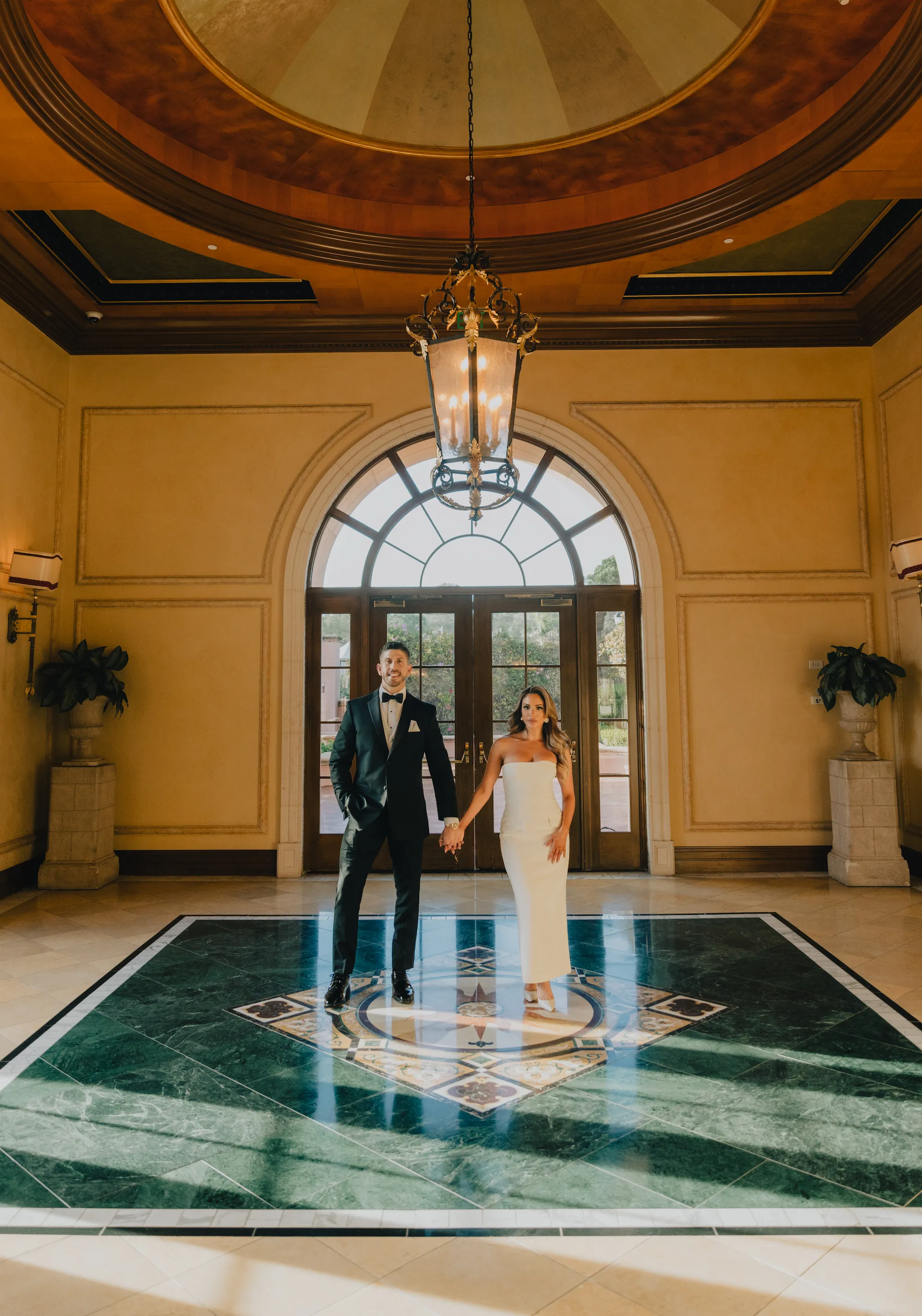 engagement photo outfits at Fairmont Grand Del Mar San Diego, couple in black tuxedo and white strapless dress standing in the grand hotel lobby with marble floors and a chandelier
