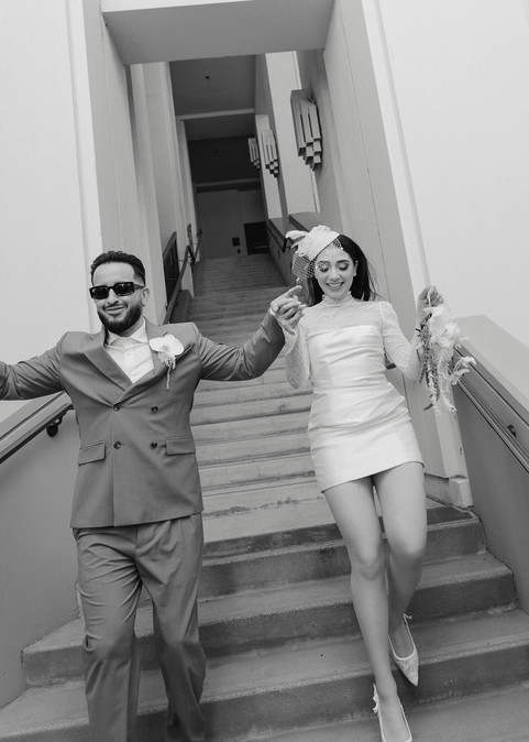 Black and white photo of couple walking down the interior stairwell at Beverly Hills City Hall after their courthouse wedding, groom in olive double-breasted suit and sunglasses, bride in short lace turtleneck dress and birdcage fascinator holding bouquet